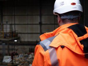 A male operative wearing a hard hat and orange high visibility jacket looks onto a waste site.